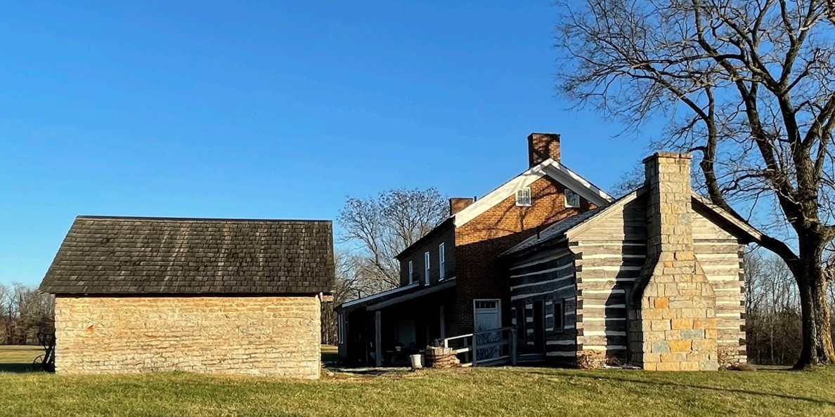 The John T. Wilson Homestead in Ohio served as a stop on the Underground Railroad. A red brick, two story house attached to an older log cabin with chimney sits next to a smaller brick barn. There are no other buildings in the photo. There are some trees and a bright blue sky.