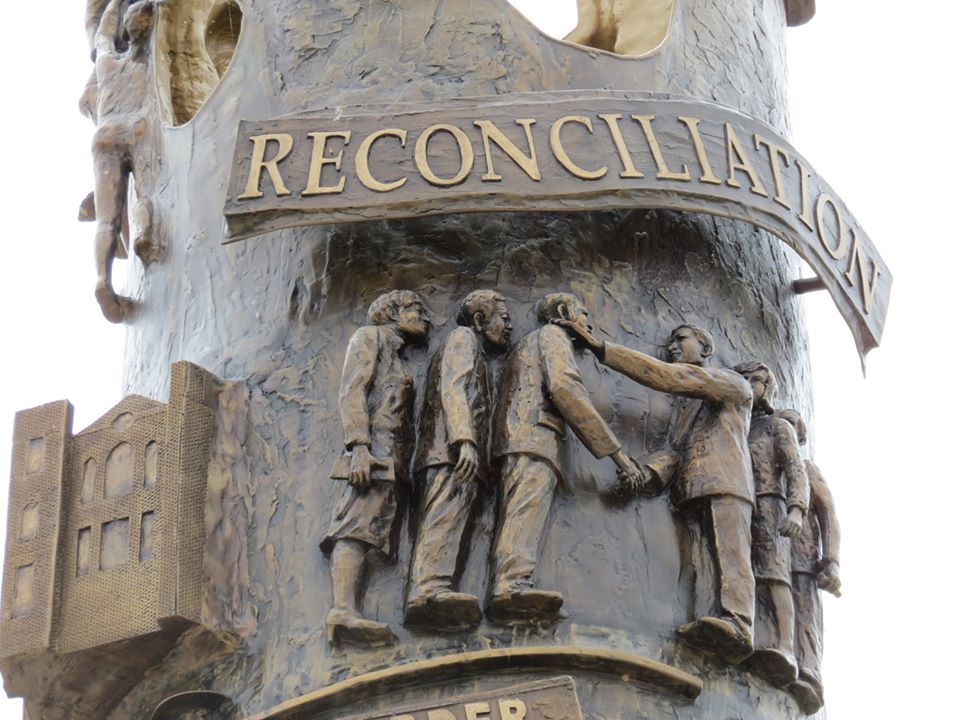 Close up of the dark bronze Reconciliation Tower. Two groups of men are depicted shaking hands below the word "Reconciliation".