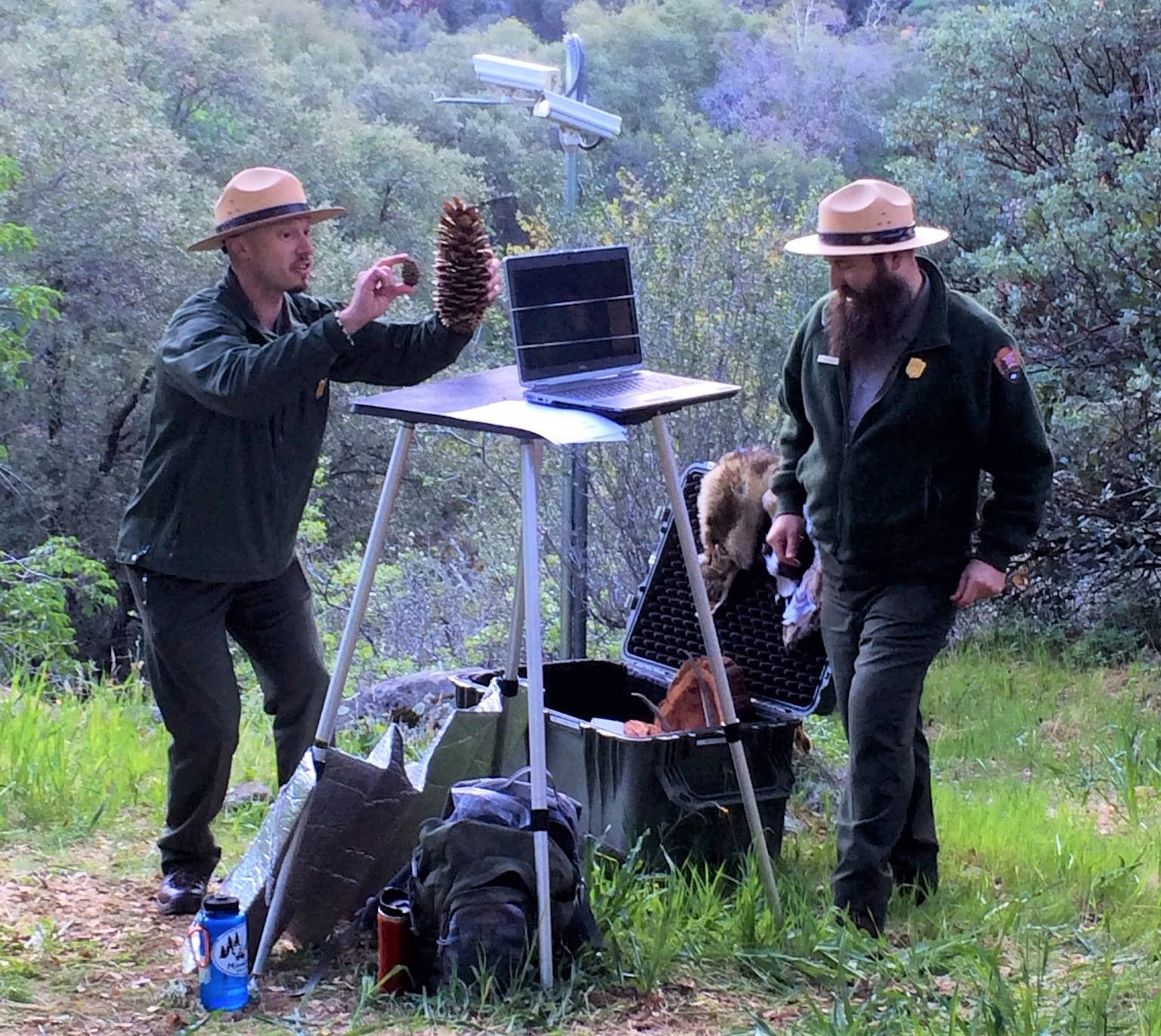 Rangers presenting a virtual education program. Ranger in front of monitor holding a small pinecone and a large pinecone with another ranger nearby.