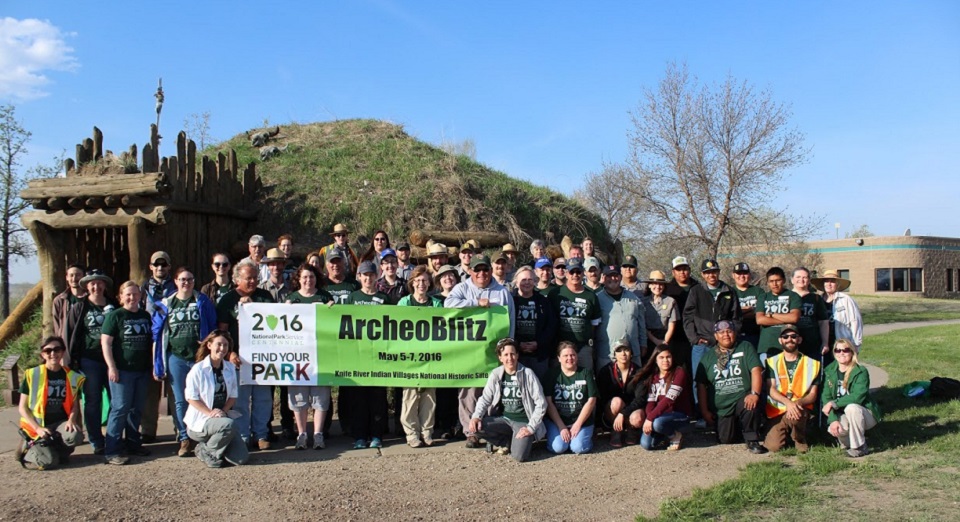 Group of people standing in front of an earthlodge holding an "Archaeoblitz" sign