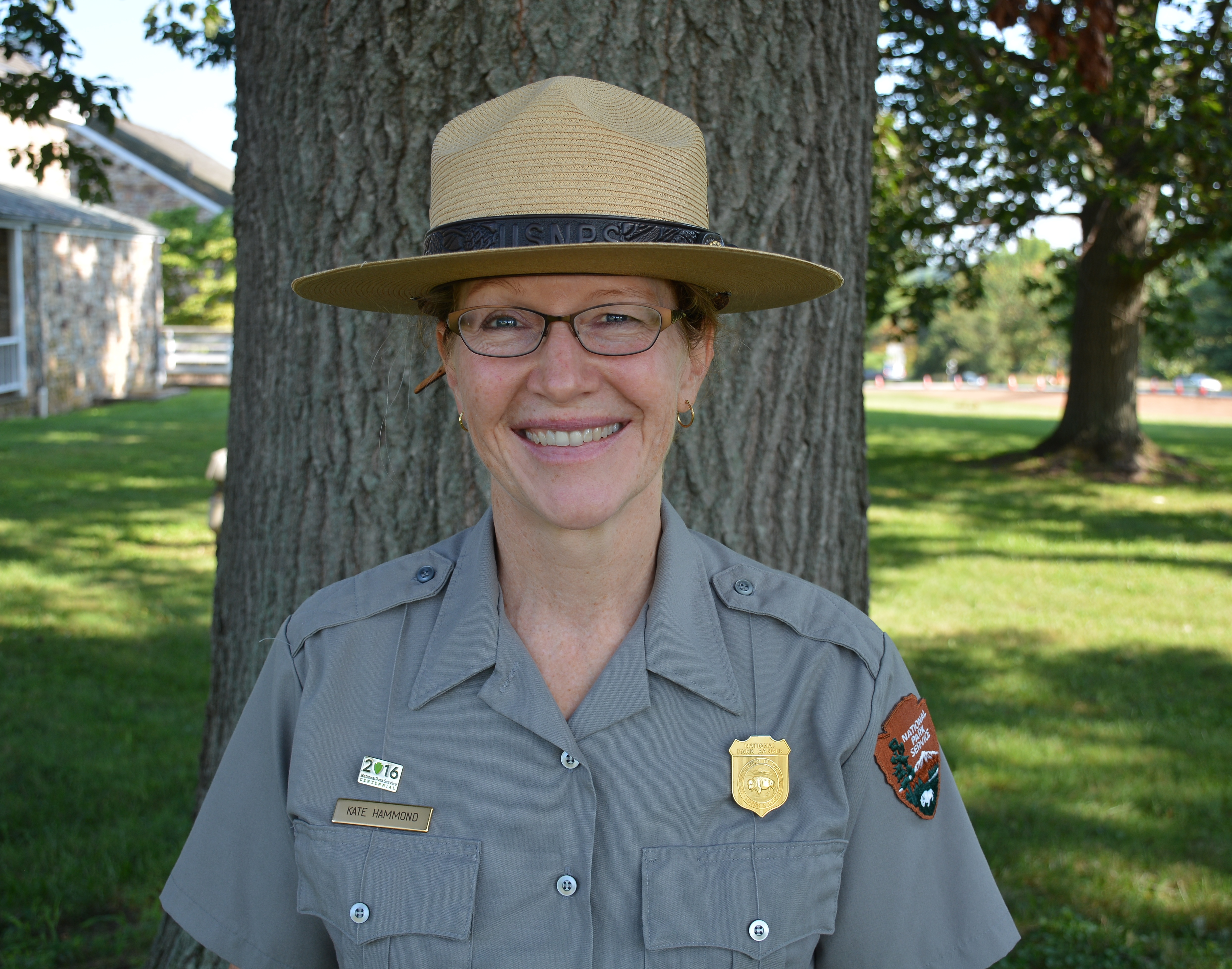 Kate Hammond A woman wearing a flat hat, gray uniform shirt, name plate and badge stands in front of a tree.