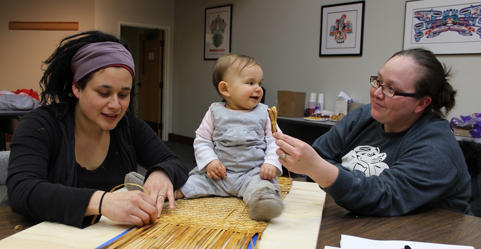 Two women demonstrating basket-weaving with a baby sitting on the flat basket