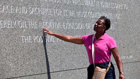 A woman in a pink shirt reaches out to a stone inscription on the exterior wall of a monument.