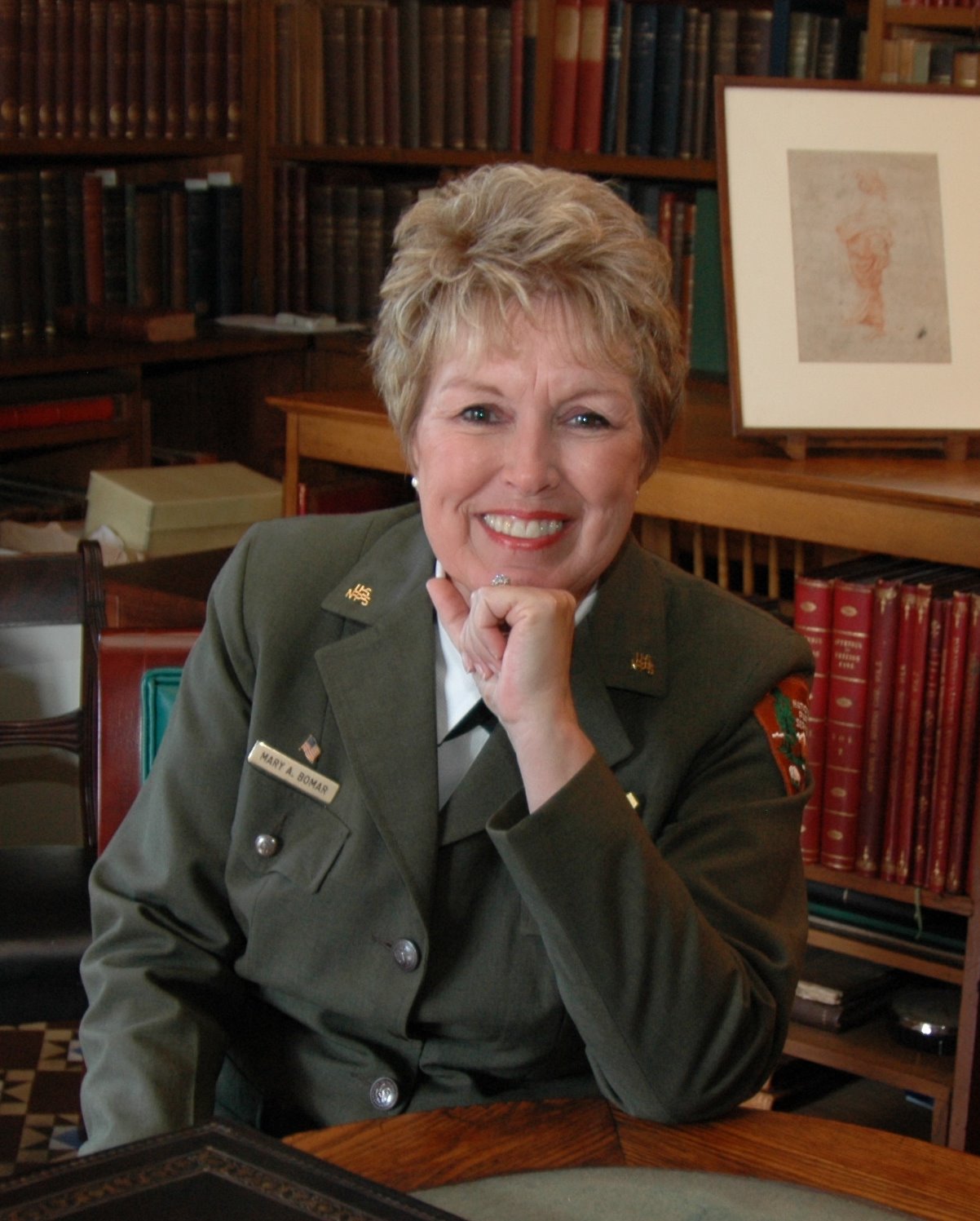 A woman in a formal green uniform rests her chin on her left hand; behind her are several shelves lined with books.