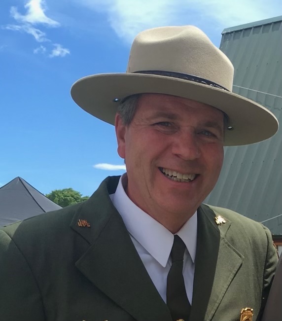 Mike Reynolds 2 A man in a green ranger jacket and tan felt flat hat stands against a background of two structures and a blue sky.