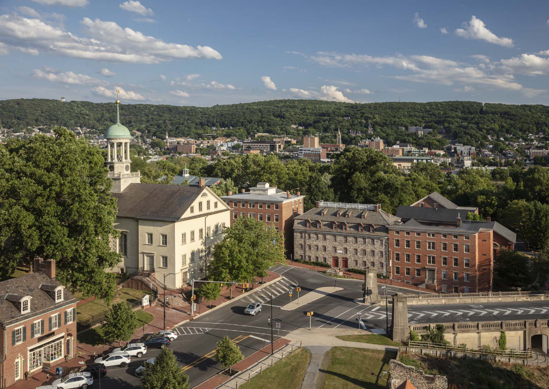 Aerial shot of the Historic Moravian Bethlehem District in Bethlehem, Pennsylvania showing several buildings around an intersecting road with green trees and a hill in the background with a blue sky and clouds.