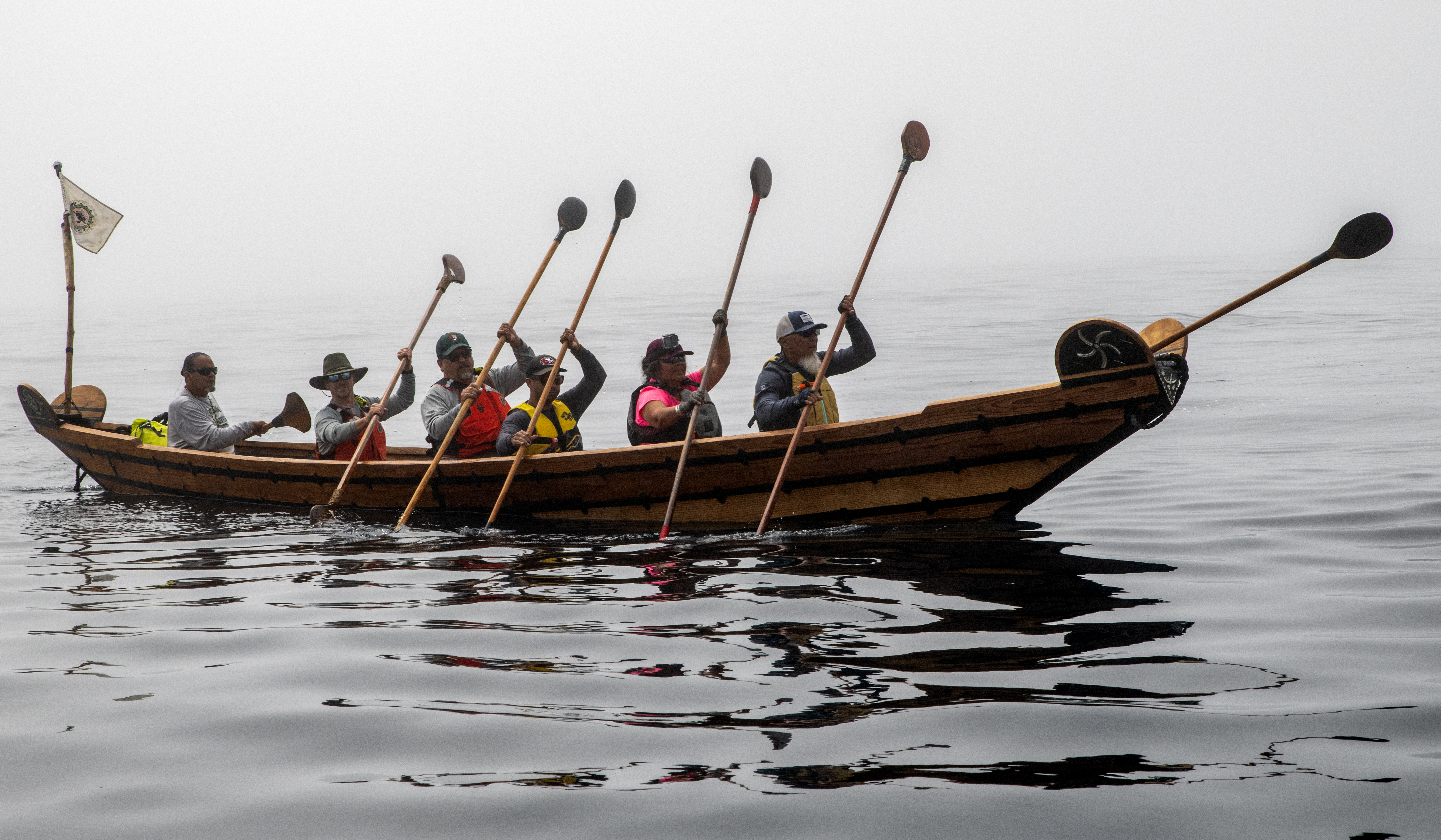 Muptami Crossing A wooden boat, with a small flag flying at the back, holds 6 people who raise their paddles in unison.