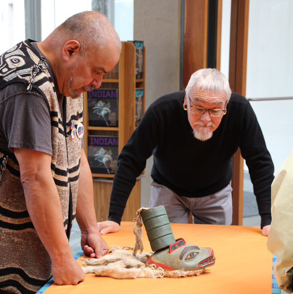 Harold Jacobs, Central Council of the Tlingit & Haida Indian Tribes of Alaska, and Joe Watkins, NPS, with Tlingit clan hat at a repatriation ceremony in 2018. NPS photo. Two specialists stand over a table looking at a traditional hat.