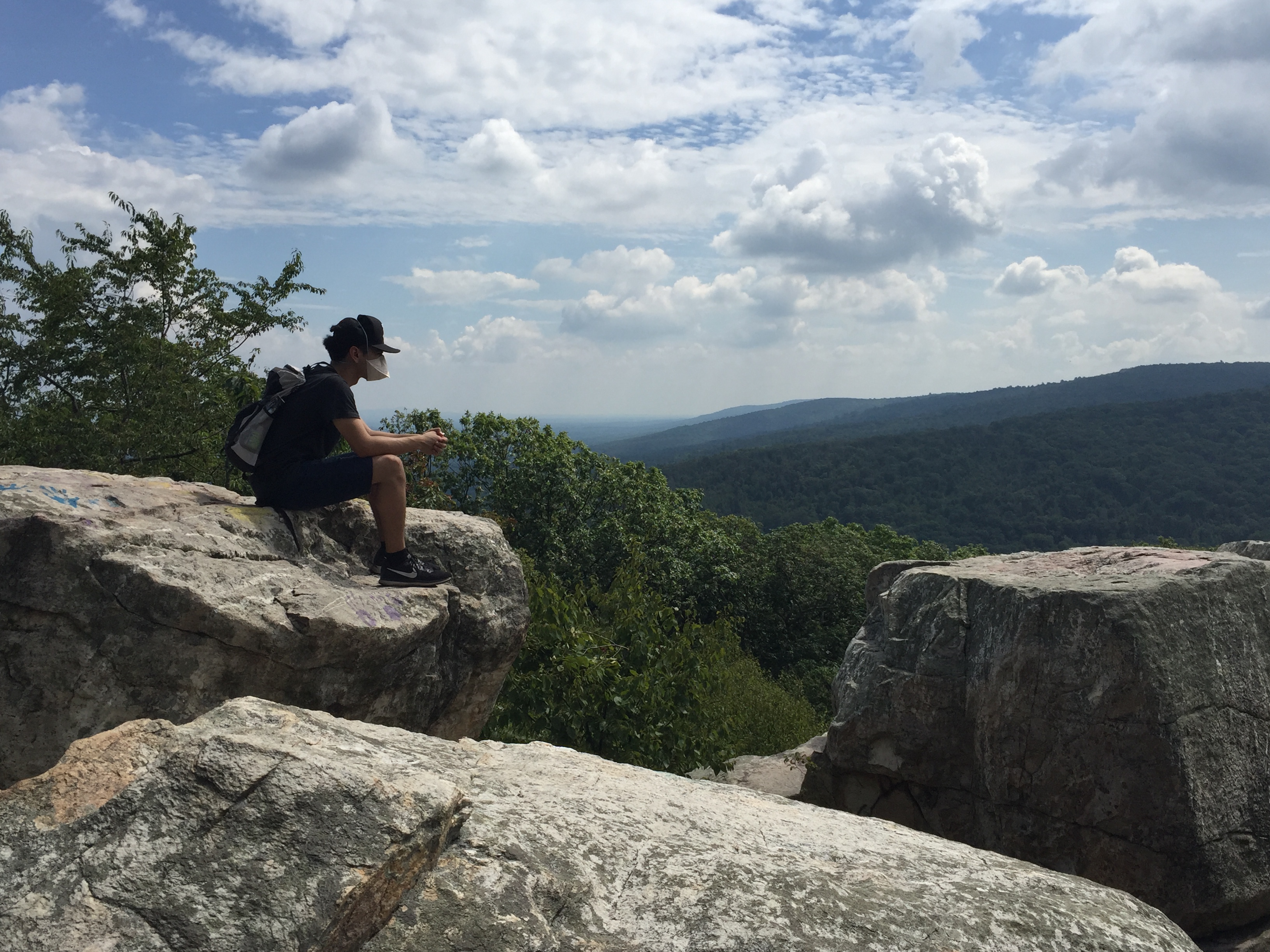 A person wearing a face mask, baseball cap and backpack sitting on a rock overlooking forested hills on a cloudy day.