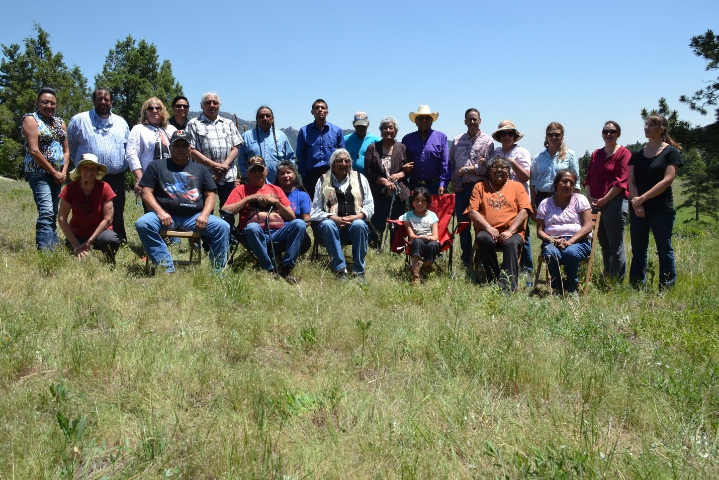 Two rows of people posed for a photo in a grassy field with the front row sitting in chairs or kneeling in front of those that are standing.