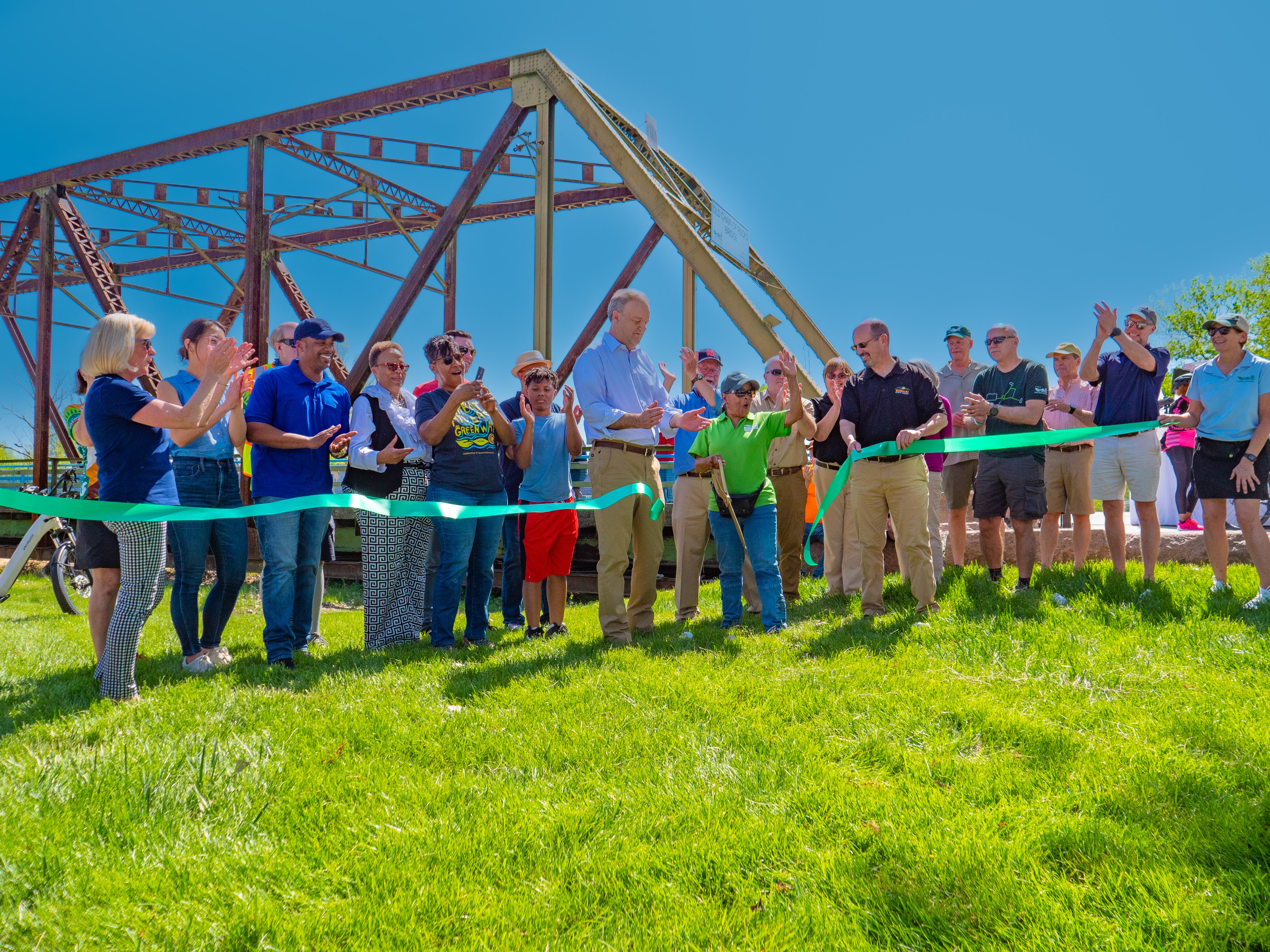 Chain of Rocks Park (MO) Ribbon Cutting People line up on a bright sunny day to cut a long green ribbon, symbolizing the opening of the park leading to a large brown metal bridge in the foreground