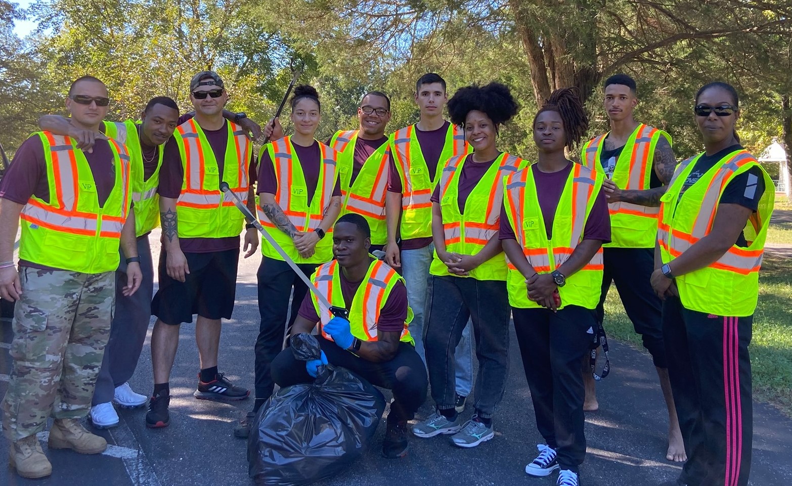 A group of 11 people in bright yellow vests pose for a photo outside along with a bag of trash they collected.