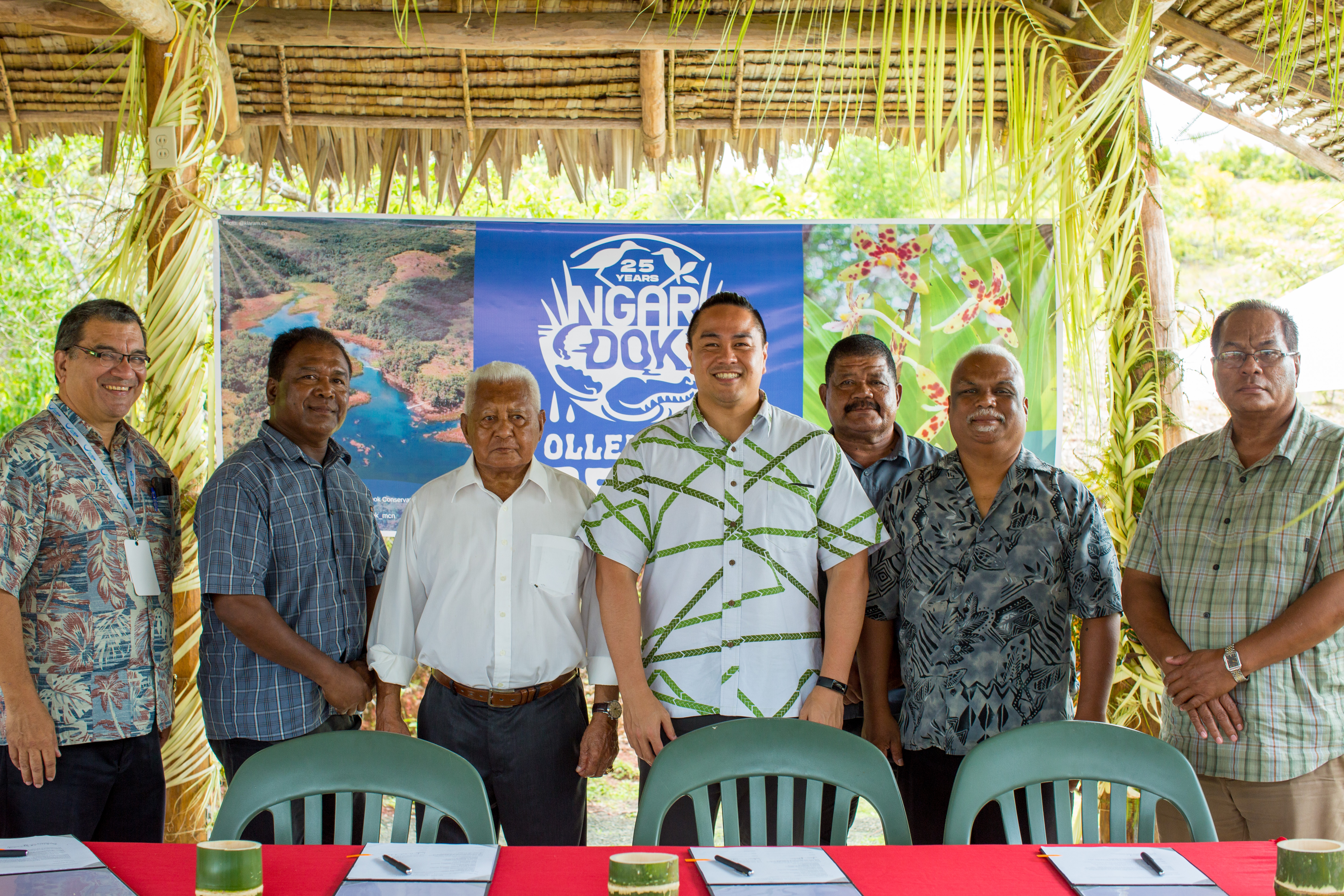 Seven men stand behind a table and chairs under a thatched canopy