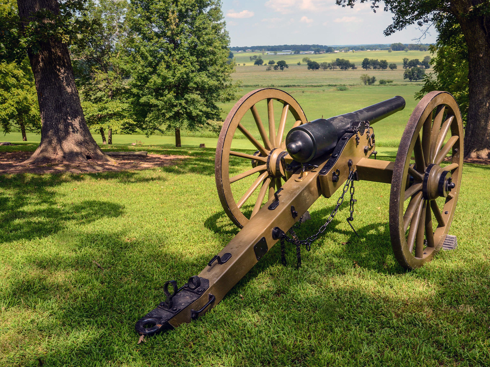 A cast iron cannon, mounted on a brown two-wheeled carriage, sits on tree-shaded hilltop overlooking green fields separated by tree lines. 