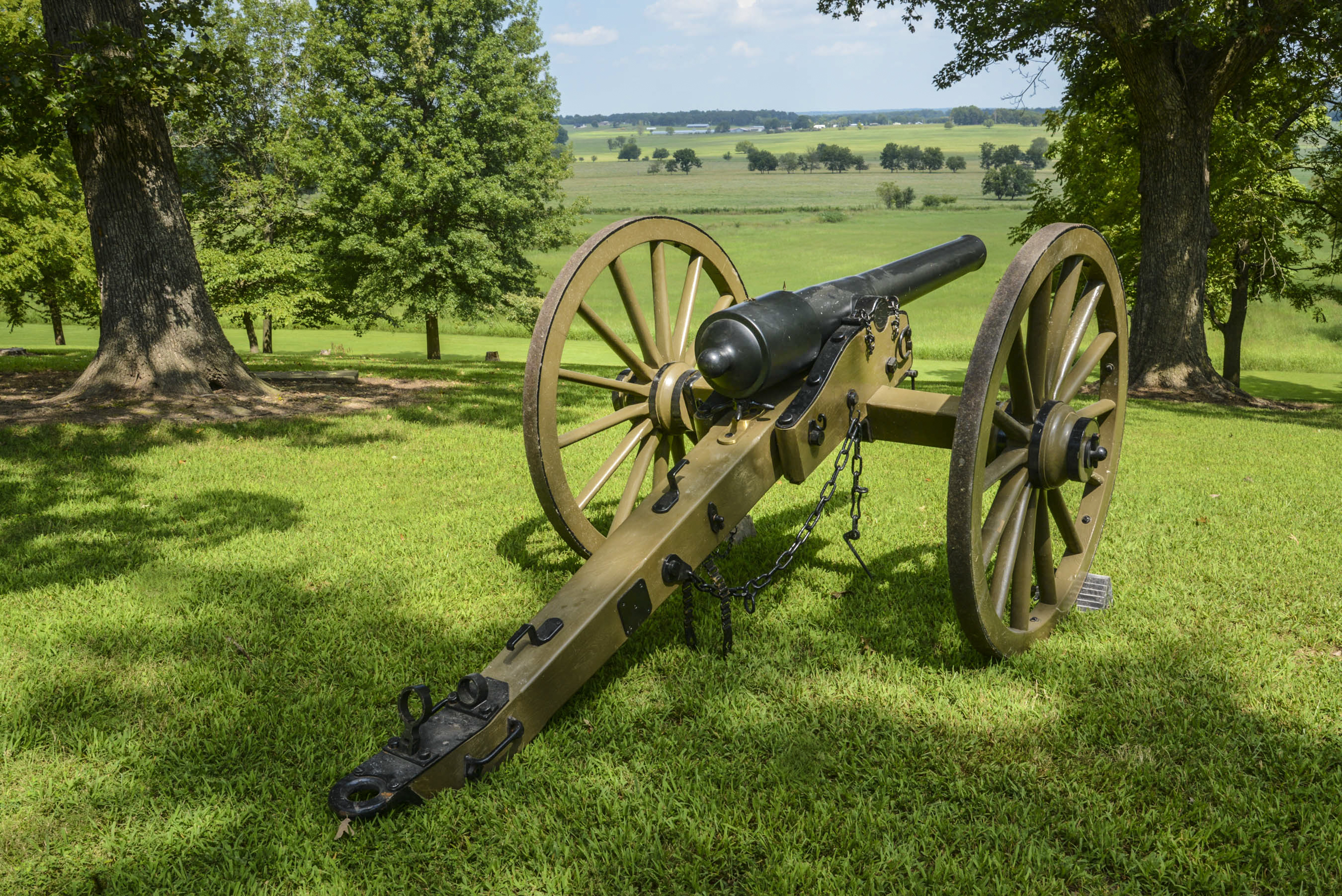 The view from Prairie Grove Battlefield State Park in Arkansas across the natural landscape now protected indefinitely. A black and brown 10-pound Parrot Rifle sits framed by two large trees to the left and right on tree-shaded, mowed grass hilltop overlooking green pastures separated by tree lines and distant buildings.