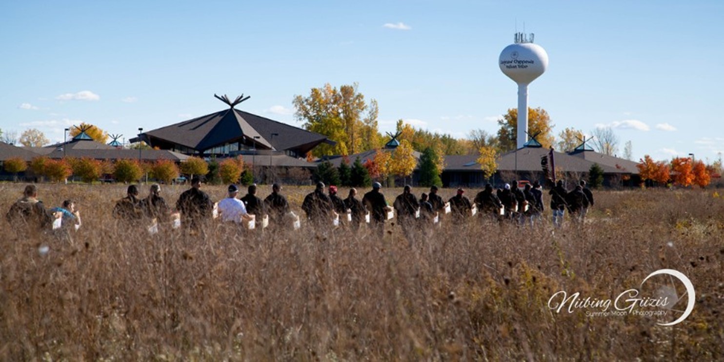 The Men’s Society of the Saginaw Chippewa Indian Tribe of Michigan carry boxes that hold 124 ancestors and 219 associated funerary objects to the Nibokaan (Place of Sleeping) Cemetery for reburial during a Recommitment to the Earth ceremony. Individuals walk in a field fed from left to the right while carrying boxes. There are several buildings and a water tower in the background.