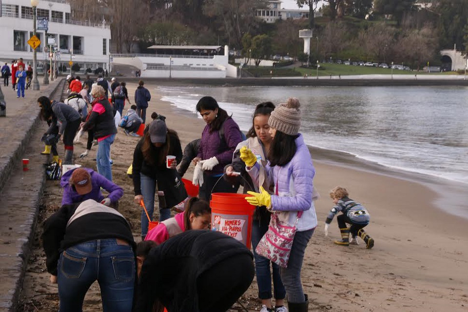 Volunteer cleanup at San Francisco Maritime National Historical Park. Group of people on a beach holding buckets. Some people bending over with arms outstretched towards the sand.
