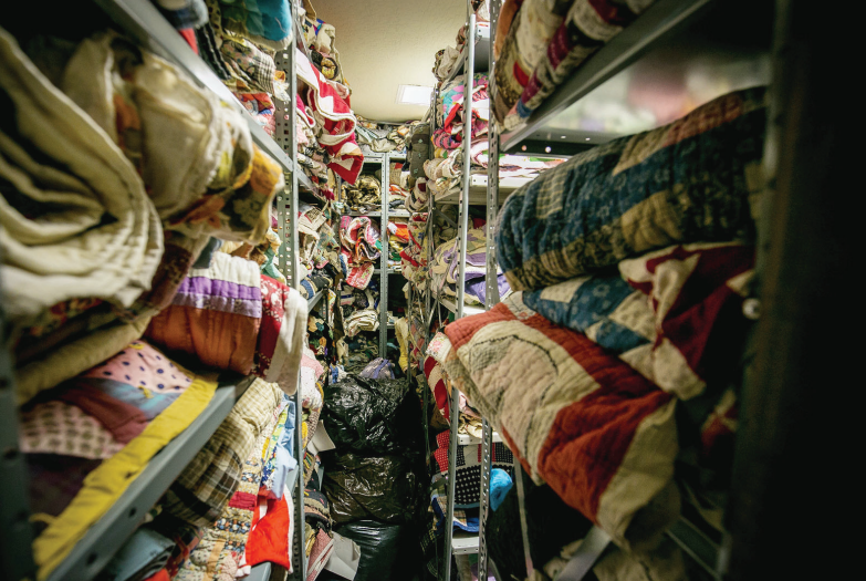 A narrow view of a column of metal shelves full of quilts of various color and designs.