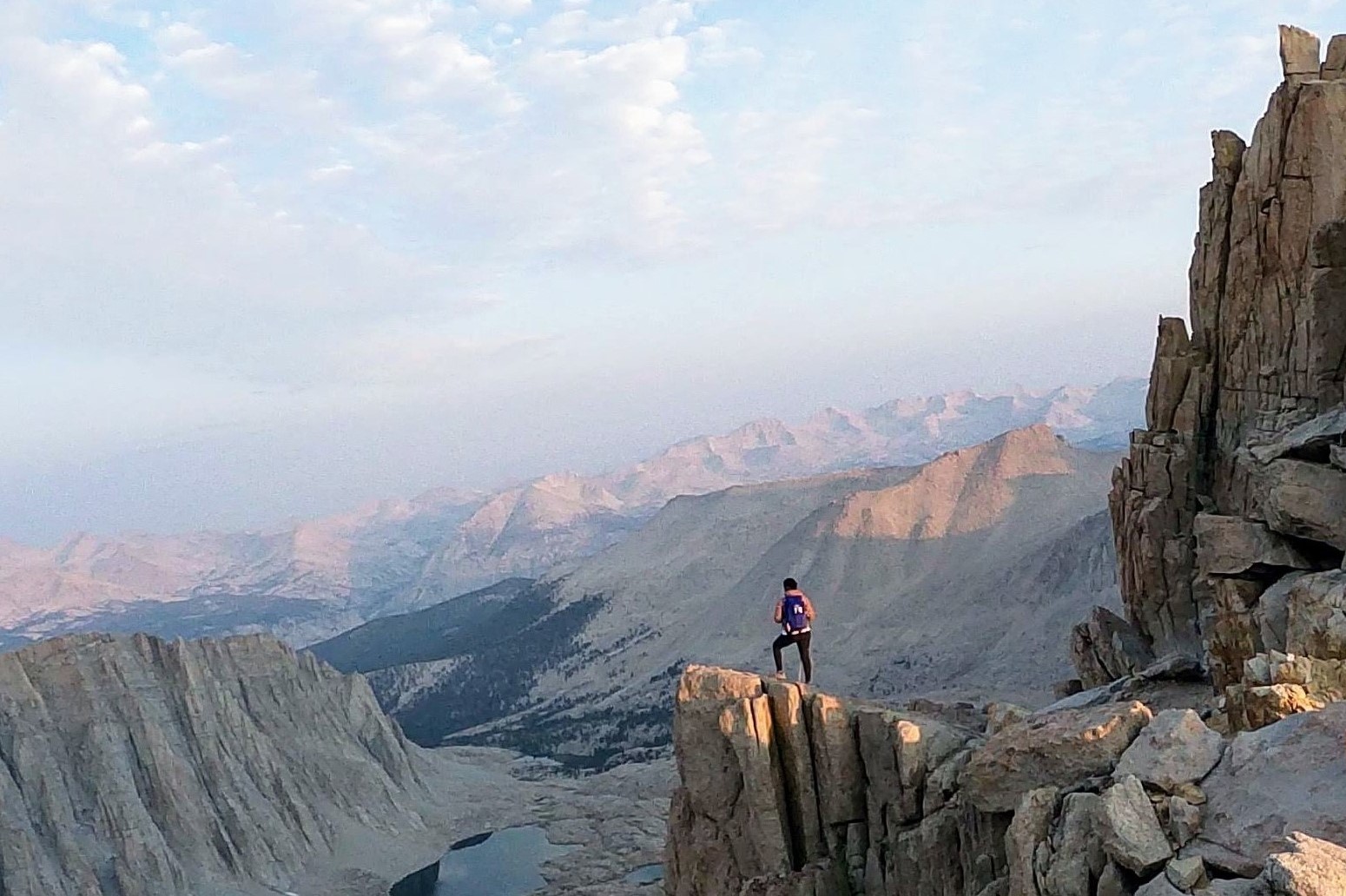 A visitor to Sequoia and Kings Canyon National Parks stands on the edge of a mountain looking over a vast landscape that includes water below and more mountains in the distance.