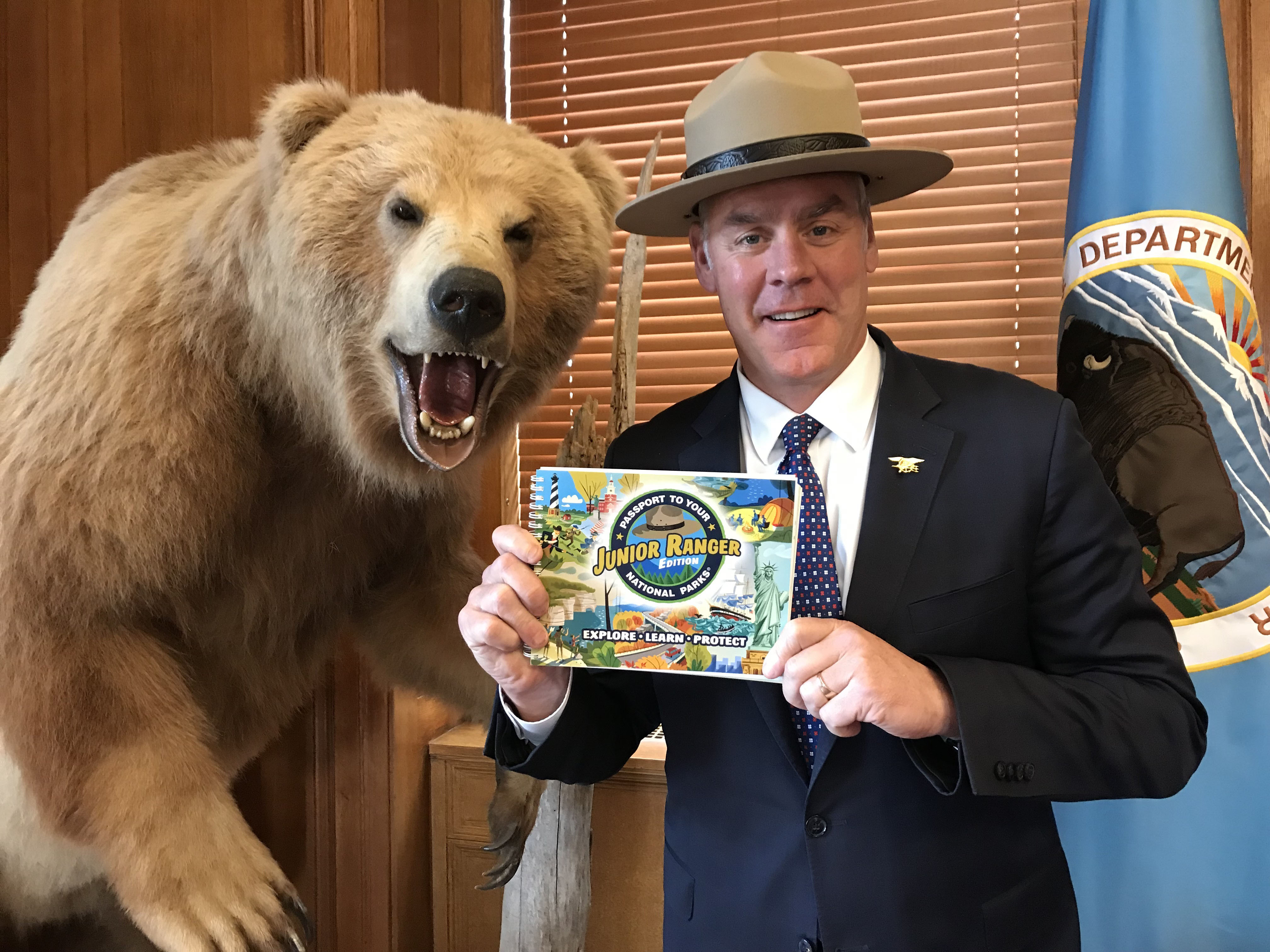 U.S. Secretary of the Interior, Ryan Zinke holding the new Junior Ranger Passport book while standing next to a stuffed bear.