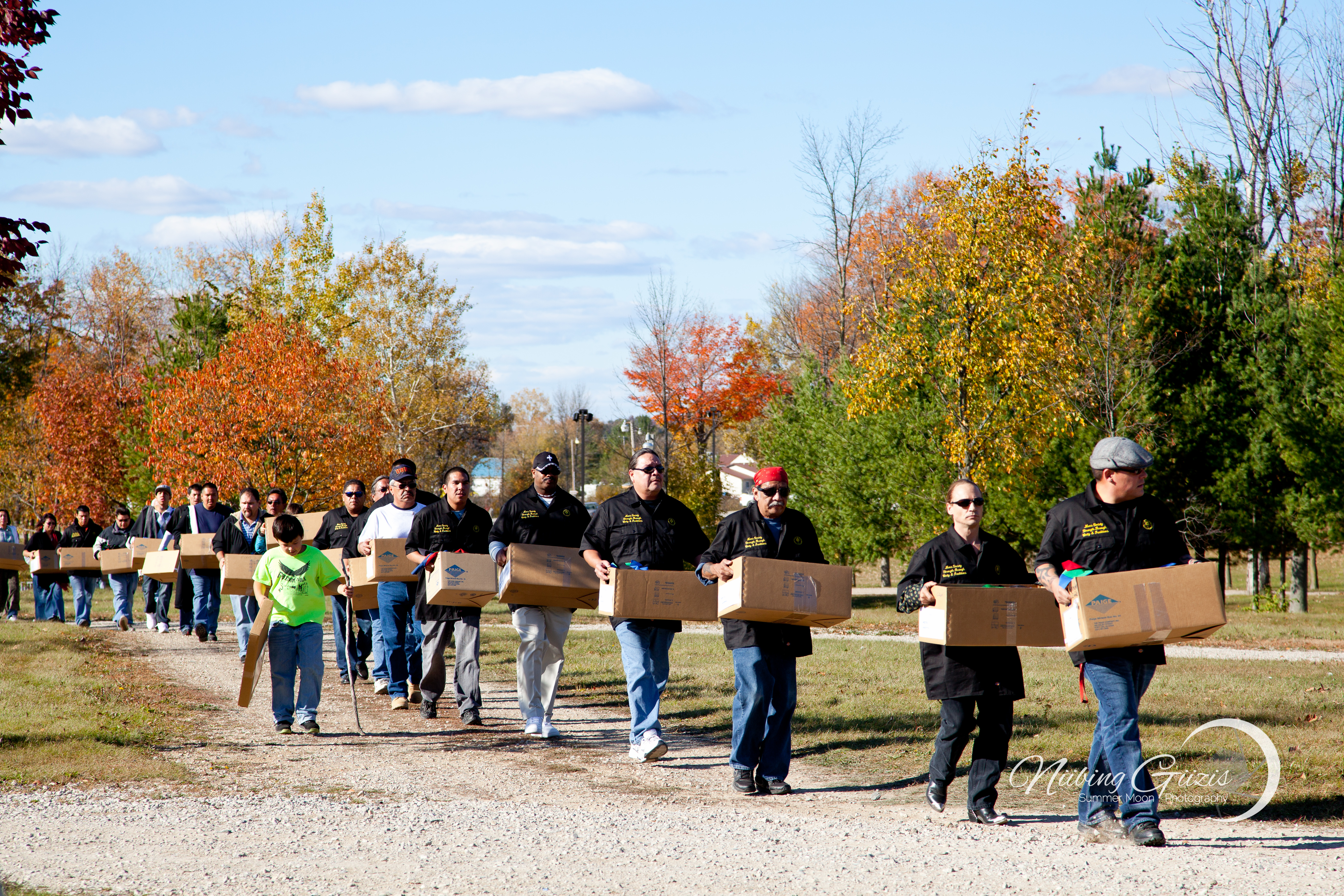 A line of people in black shirts, each carrying a cardboard box, walk single file along a trail.