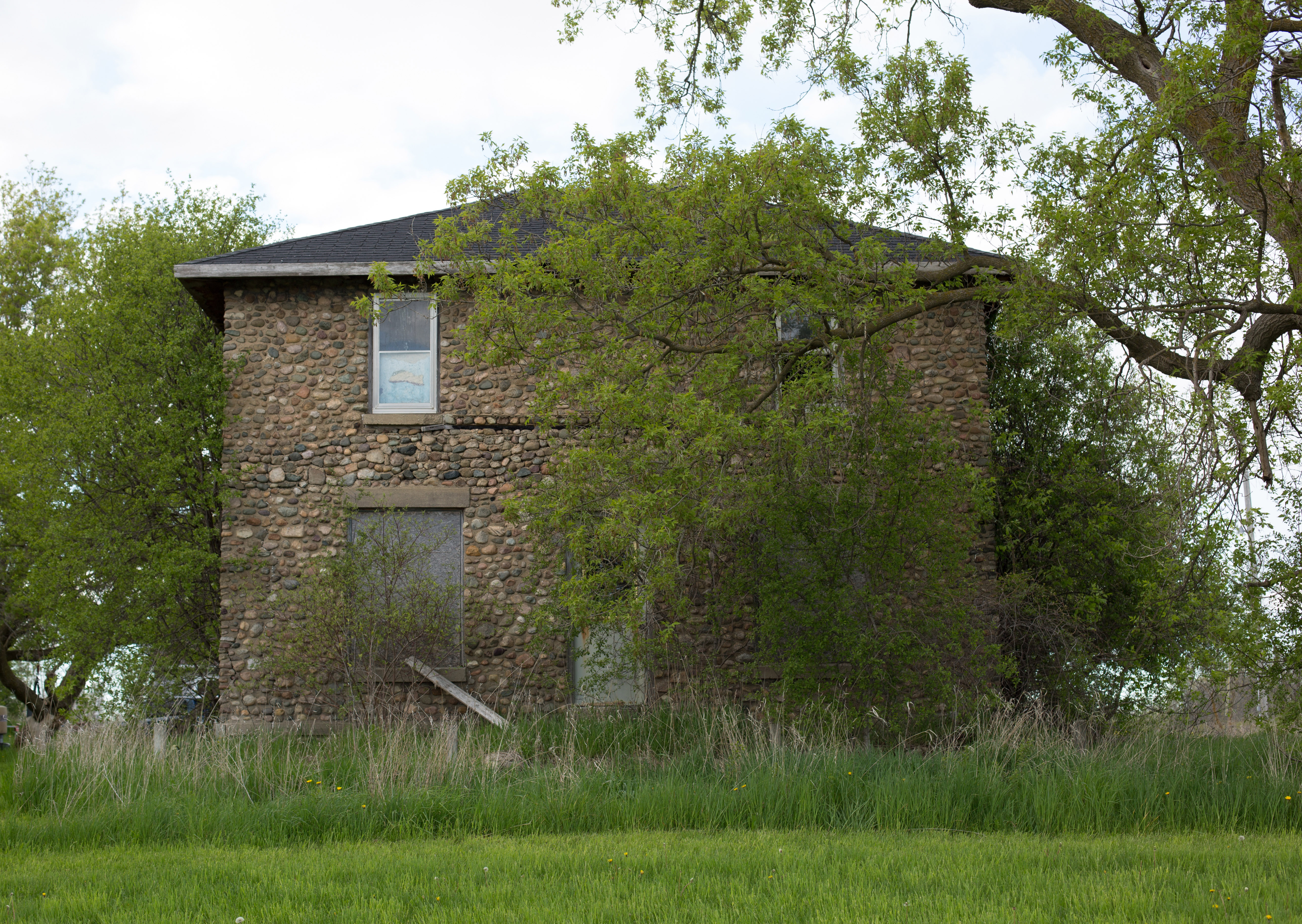 A two-story stone house with trees on either side of it, and a tall tree with a long branch reaching across it in the foreground.
