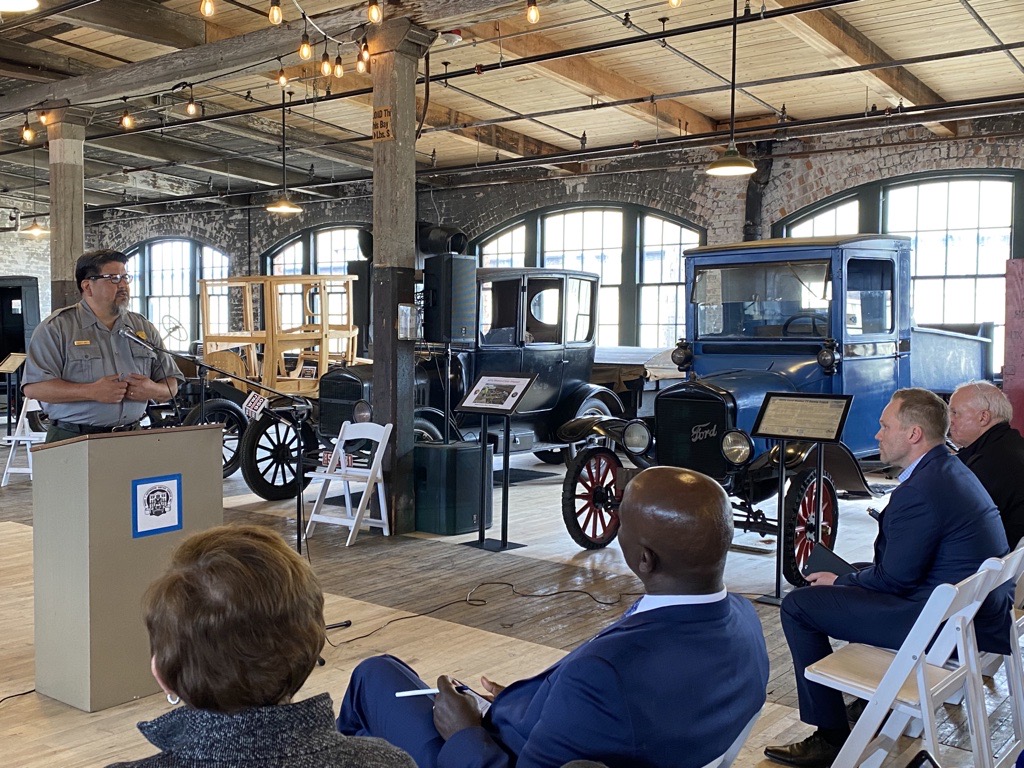 Against a backdrop of antique automobiles, National Park Service Director Chuck Sams stands behind a podium (at left) addressing a small, seated audience (at right).