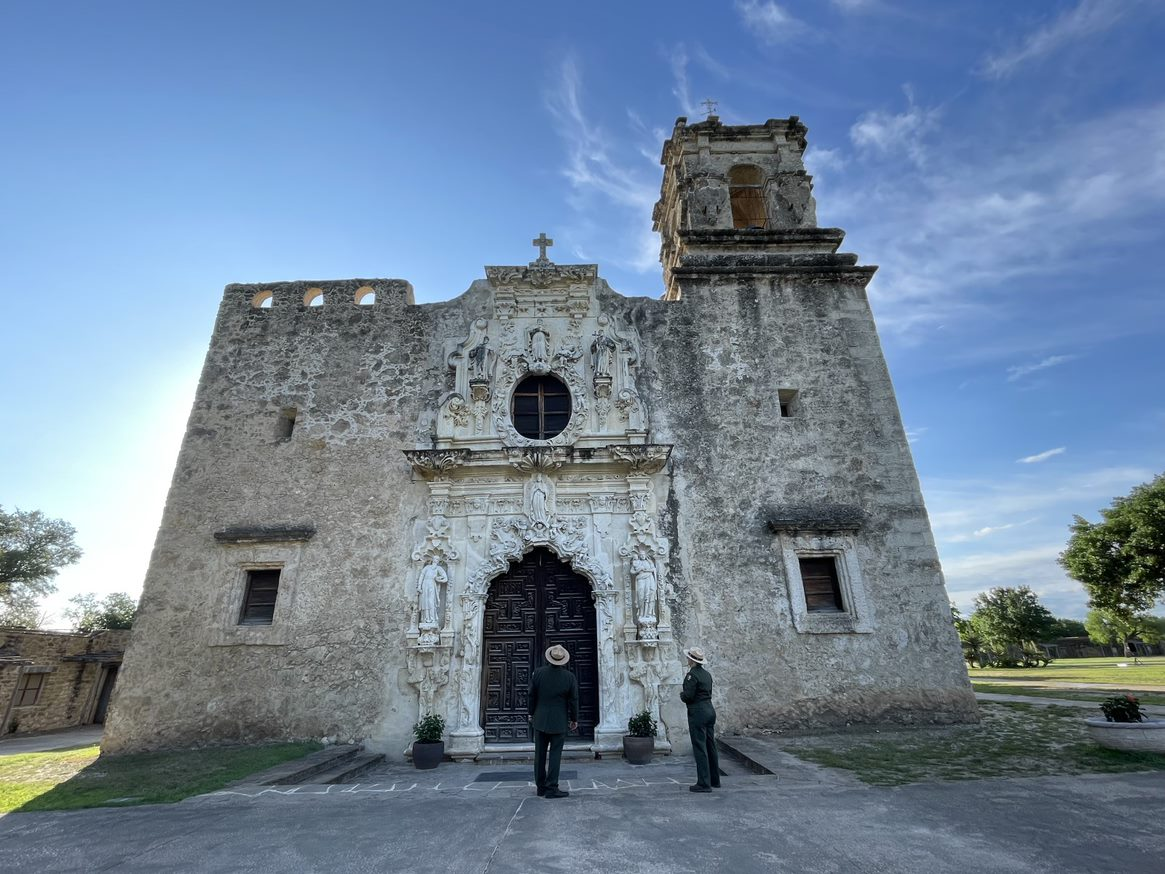 Two people, dressed in ranger uniforms, look away and up at a Spanish mission-style building.