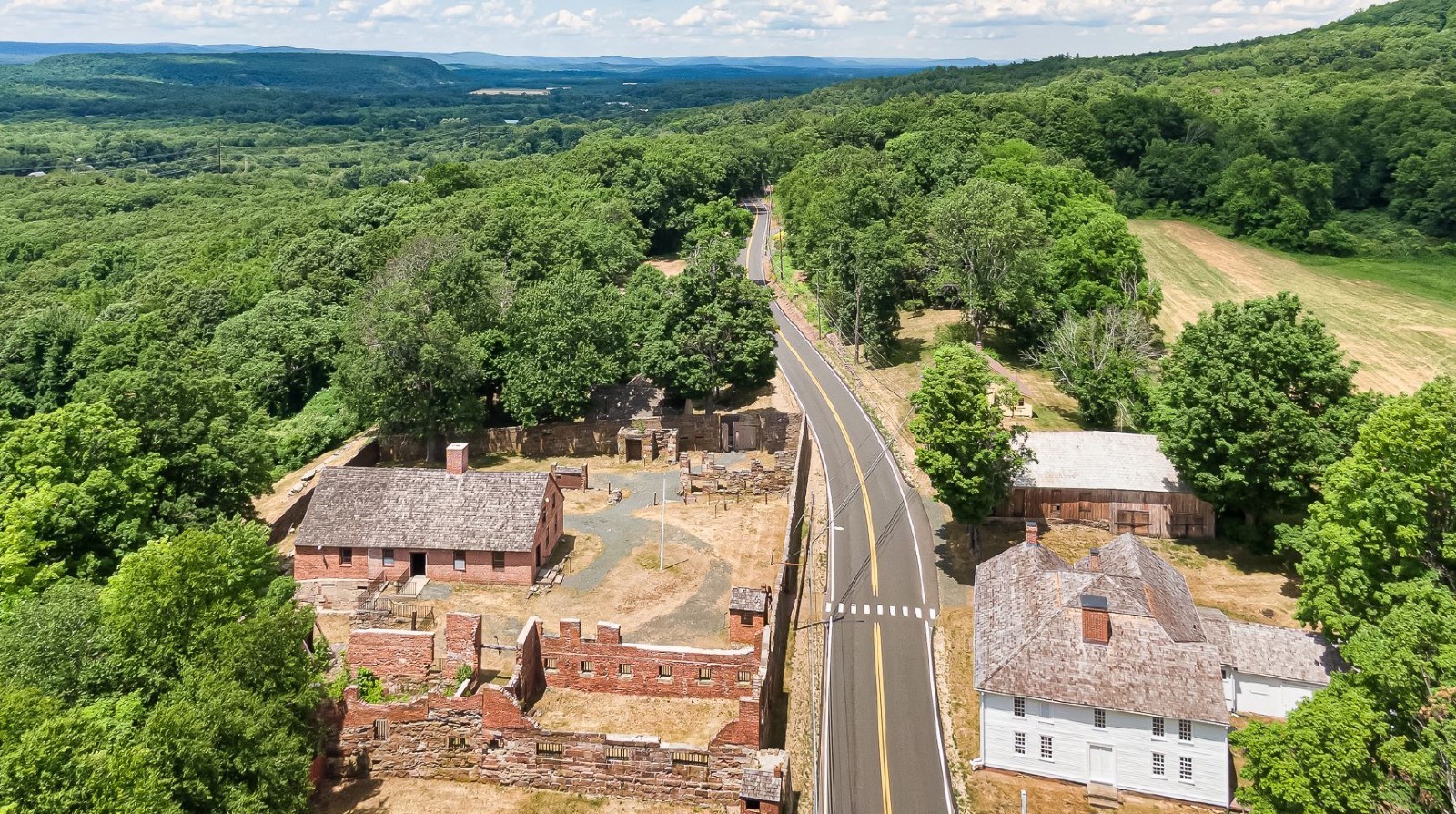 Bird's eye view of a crumbling brick structure next to a brick building surrounded by a brick wall. Across the street is two other old buildings. These structures are surrounded by trees.