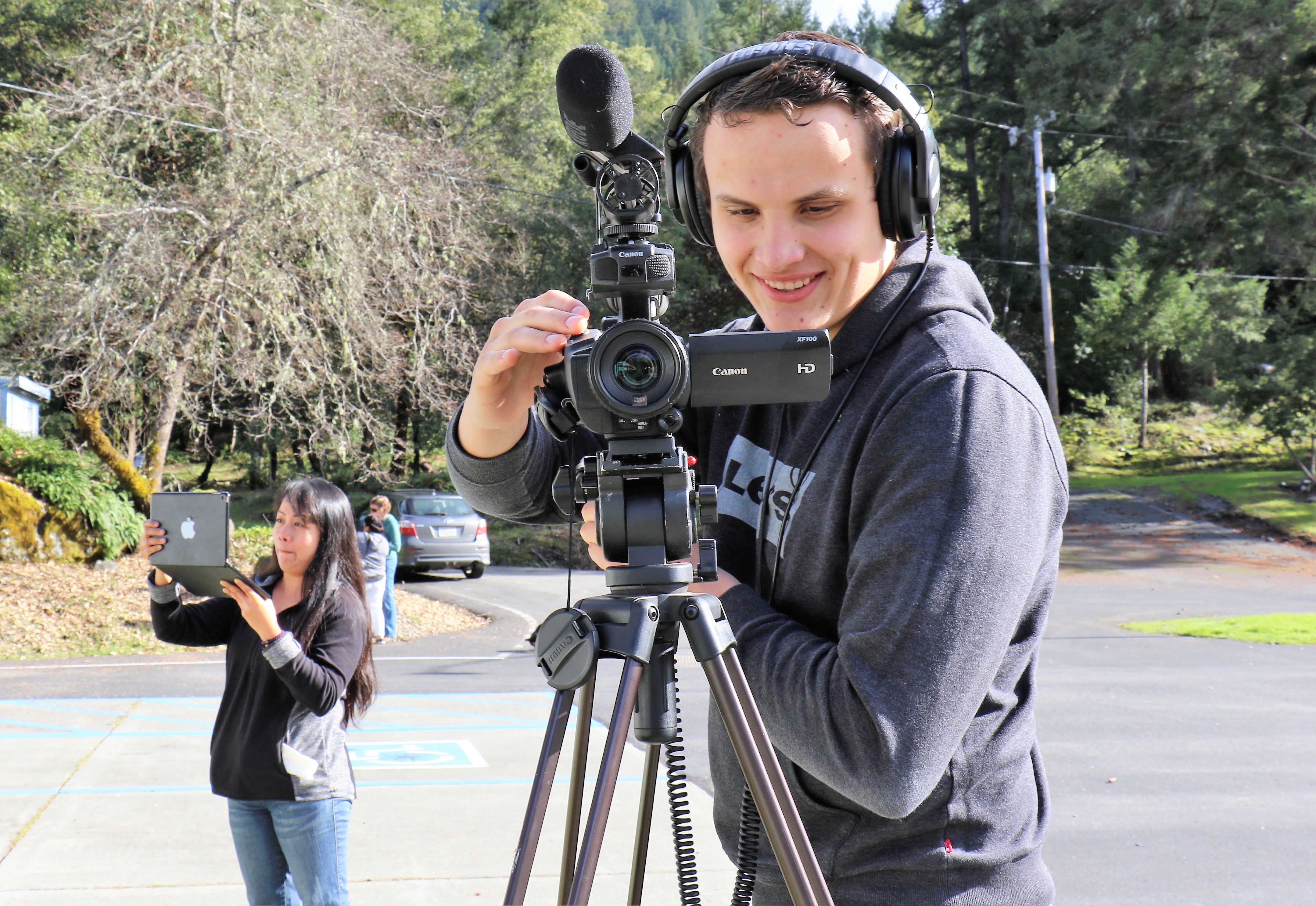 A young man wearing a black sweatshirt and headphones looks into a camera on a tripod as he smiles. A woman looking at a laptop in her hands in the background.