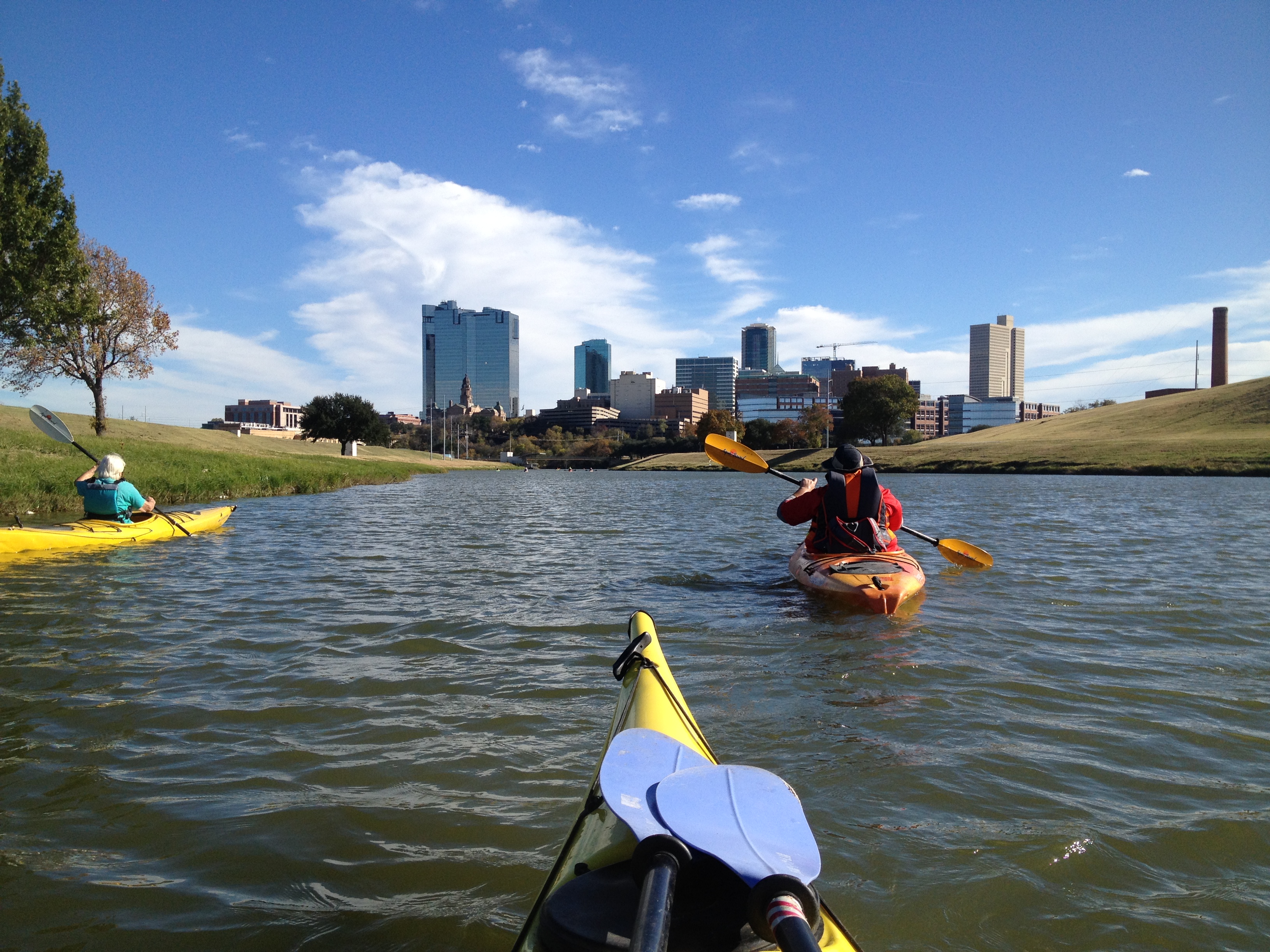 Three people kayaking on a river lined with grass and dotted by a couple of trees on a sunny day, a city with tall buildings in the distance.