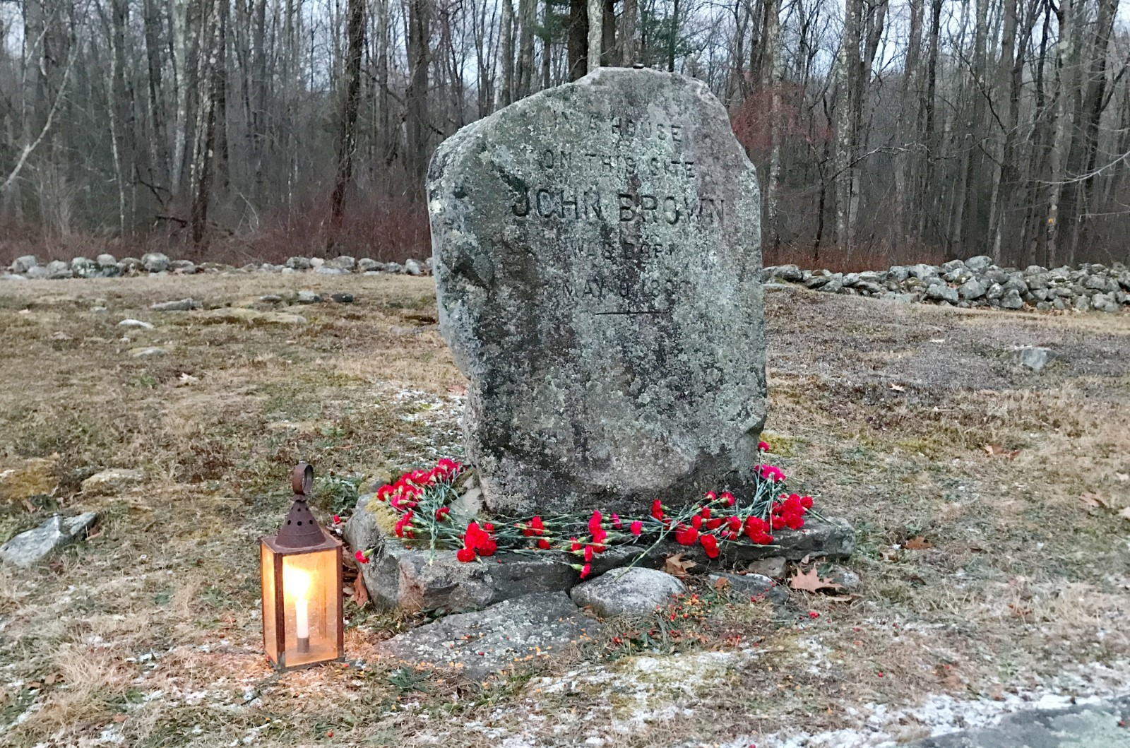 John Brown Birth Site stone marker A heavily aged and stone marker with "John Brown" carved into it. The base is surrounded by red flowers and there is a lit lantern to its left.