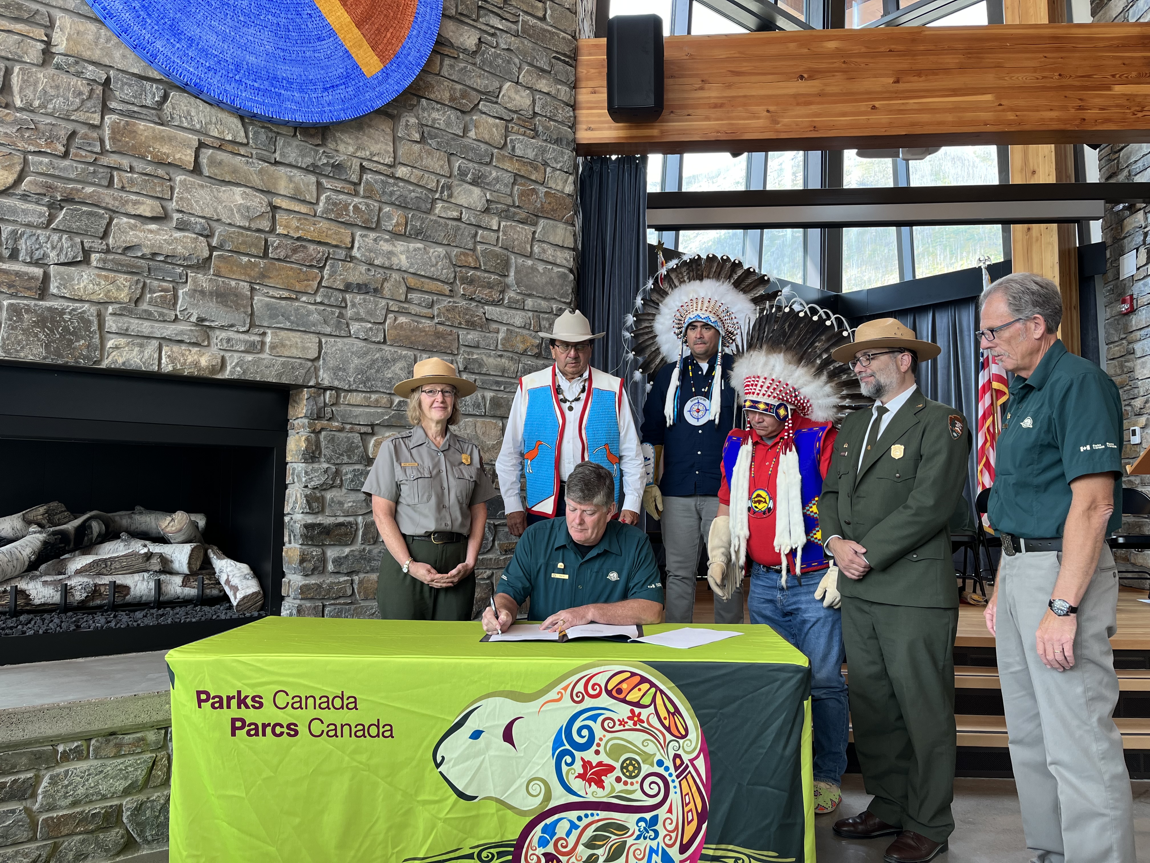 USA Canada MOU signing A man sits behind a table, with a pen and a book. There is a green tablecloth with a colorful drawing of an animal; it reads "Parks Canada Parcs Canada". Behind the man are 6 people standing.