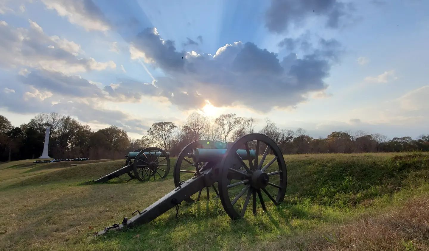 Canons set in a grassy field under an impressive sunrise peeking through clouds.