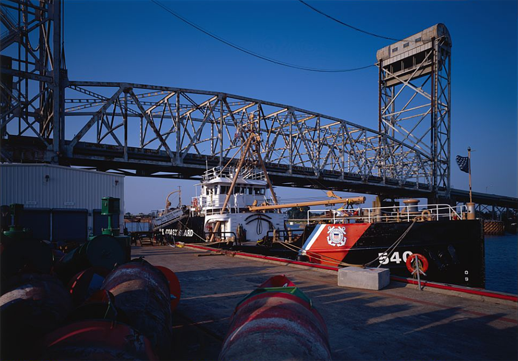 A U.S. Coast Guard Cutter is moored off a concrete pier. A bridge extends above into a blue sky.