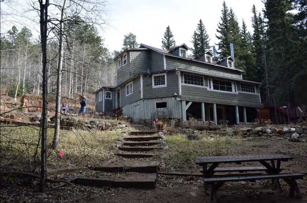 Two-story wood shingle lodge building on raised piers with an enclosed porch, located in a wooded setting with stone terracing and log fencing.