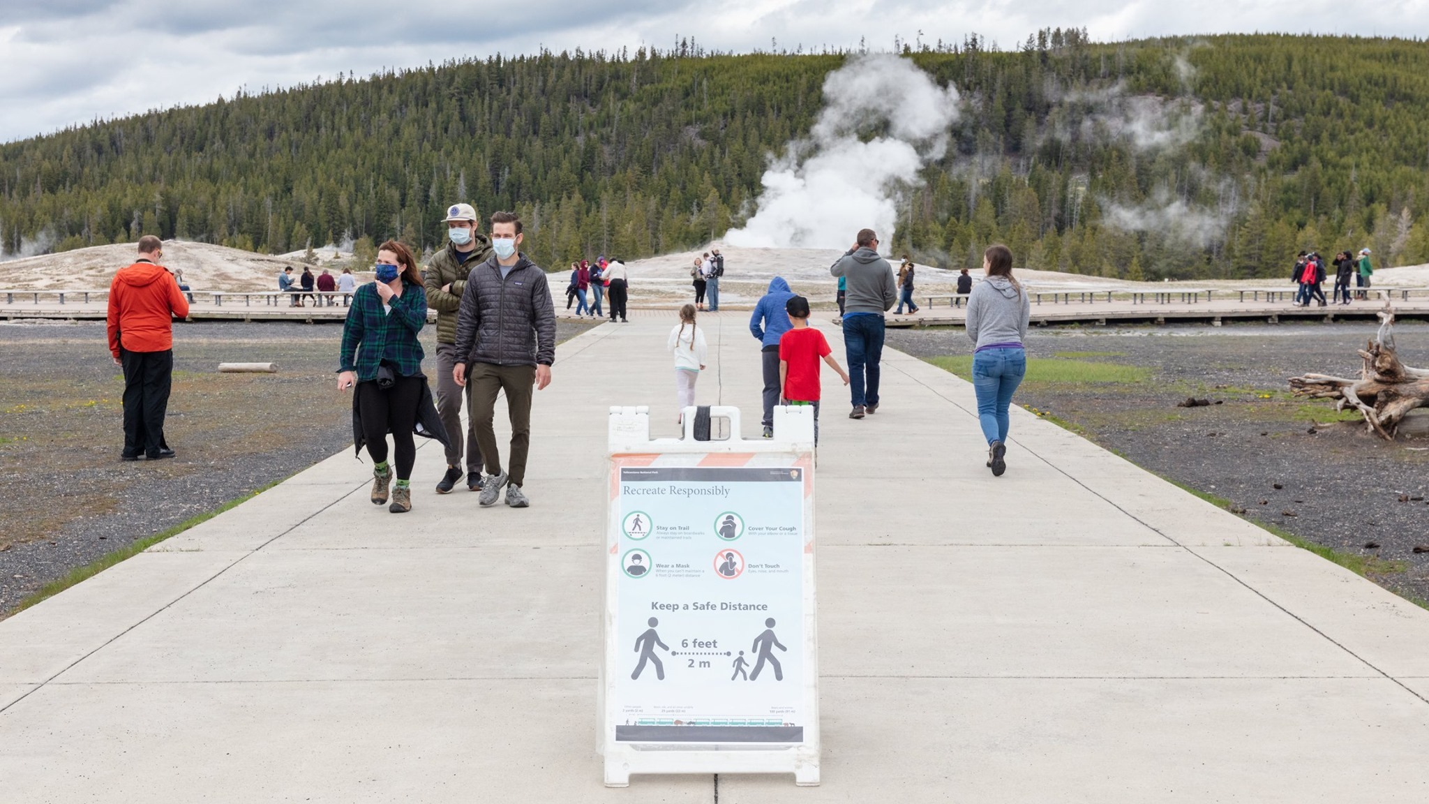 Visitors wearing face masks use a walkway that leads to a steaming geyser in the background. The groups of visitors are physically distant. In the foreground is a sign propped on the ground that reads "Recreate Responsibly. Stay on the trail."