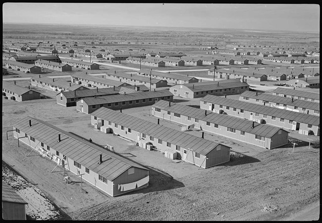 In a black and white image, a series of low-slung building stretch across a plain.