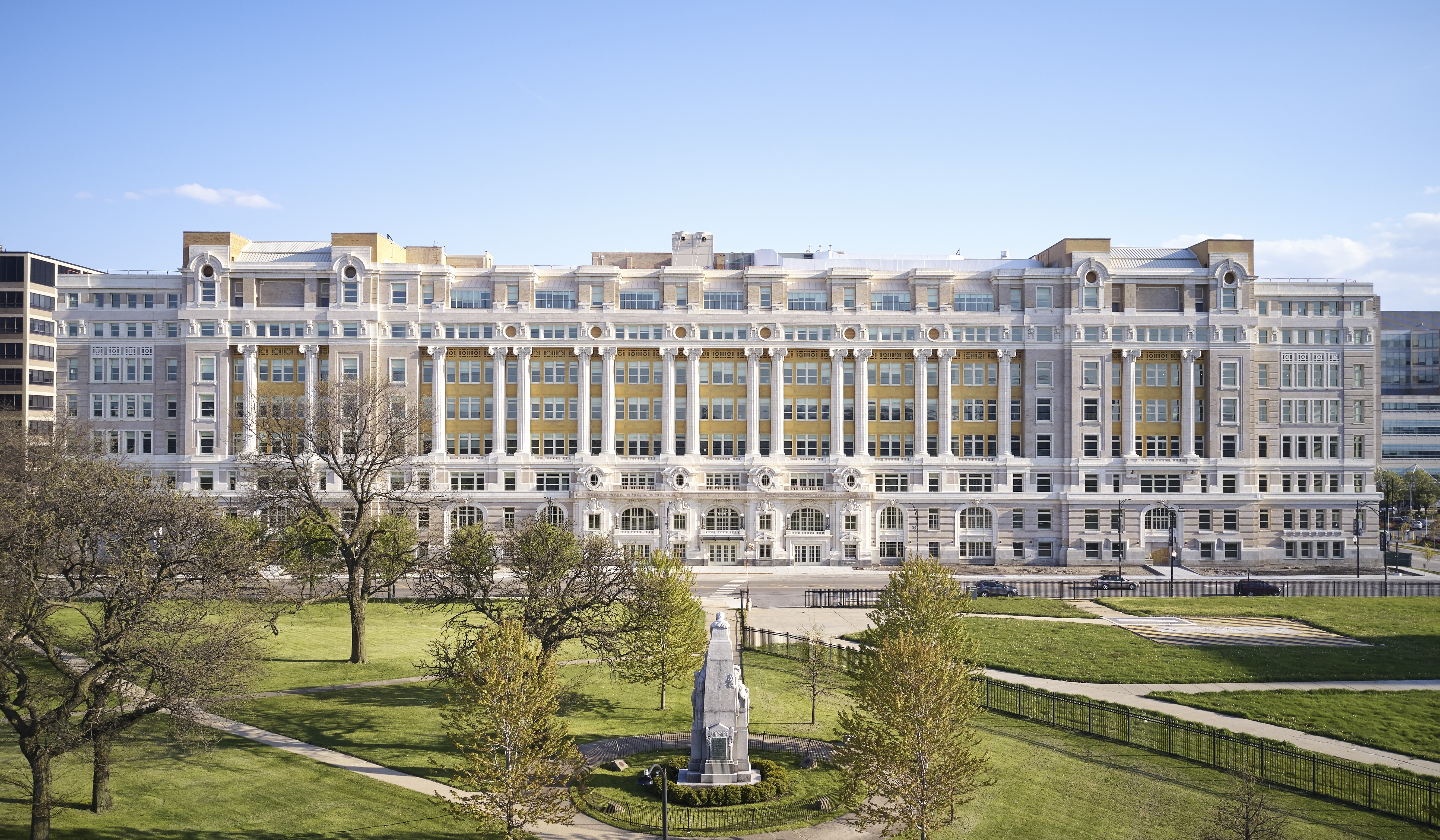 A multi-story historic building with grassy walking area in the foreground, including a statue and trees.