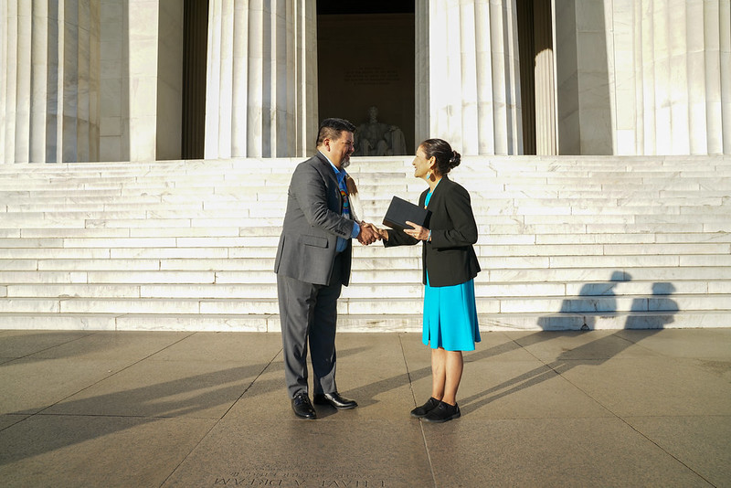 A man and a woman shake hands and smile in front of marble steps and marble columns.