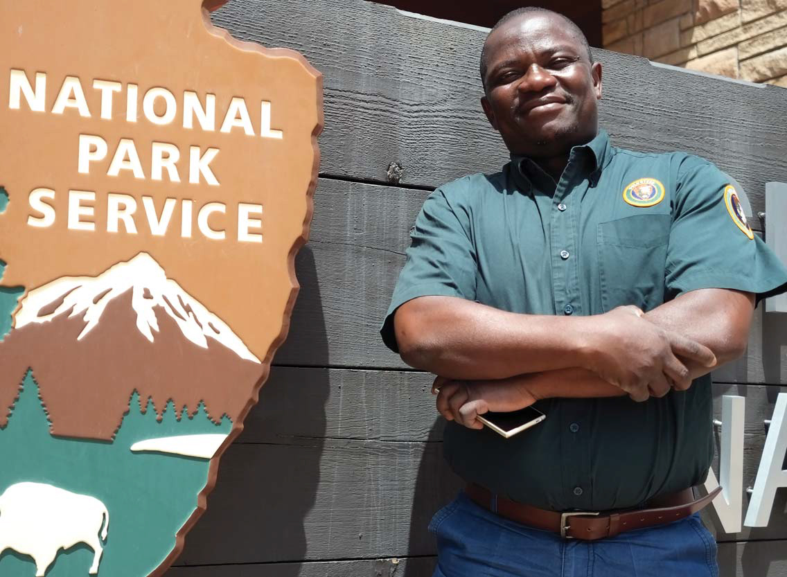 A man wearing a dark green polo with an insignia patch poses next to a large sign with the National Park Service logo on it.