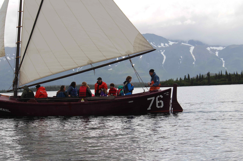 A long sailboat with 11 passengers of various ages sailing in a bay with mountains in the background.