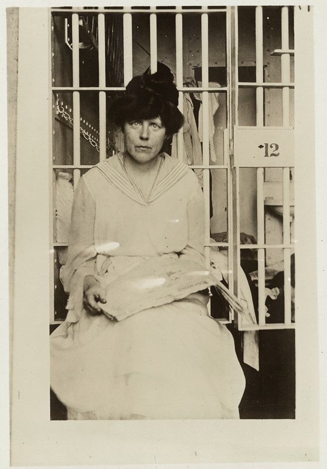 historic photo of a woman in white dress and dark hat with a jail cell door behind her