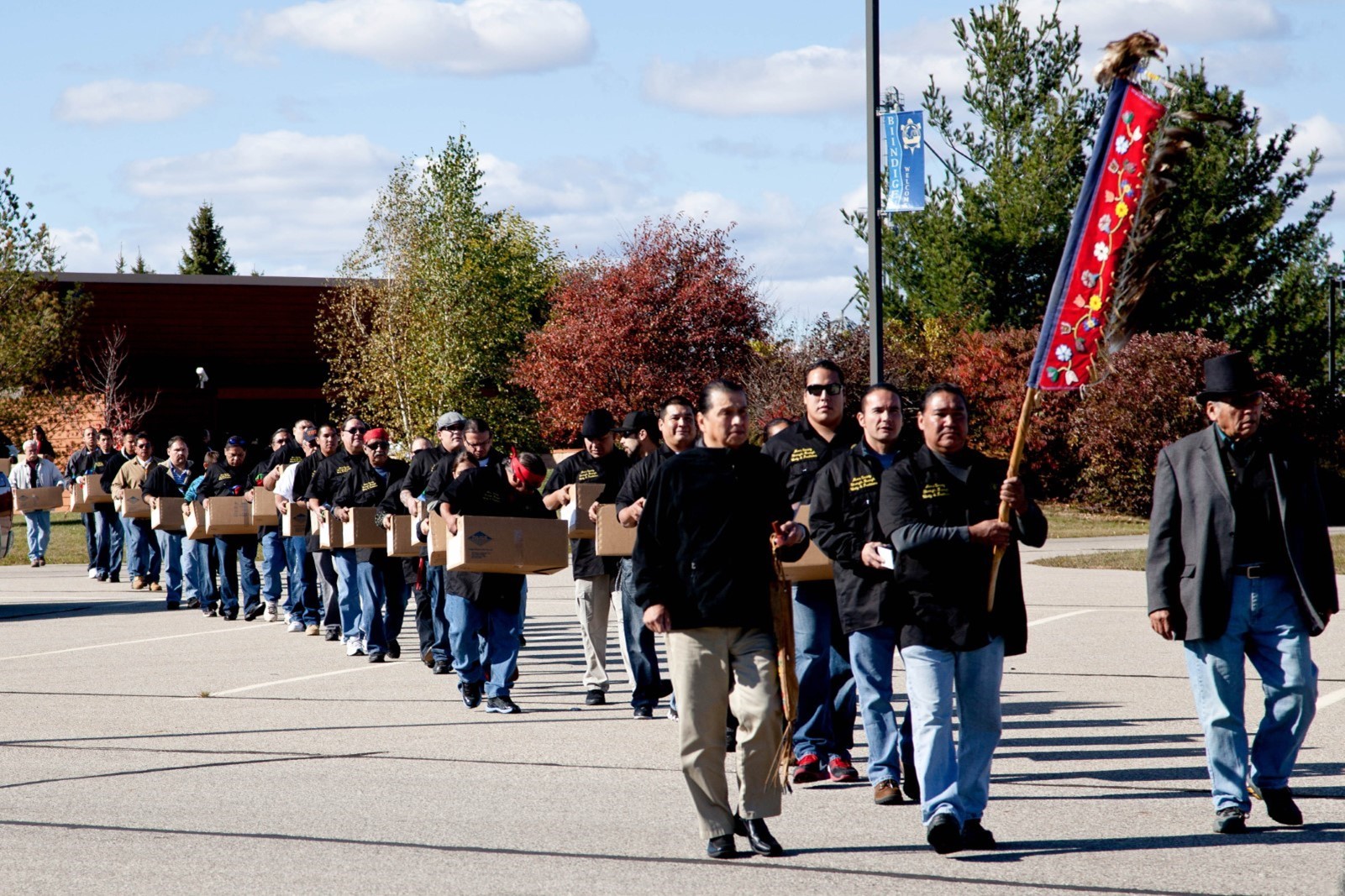 Individuals walk in a line from the left of the photo toward the right of the photo while holding boxes. There is an individual at the front of the line of people holding a ceremonial object.