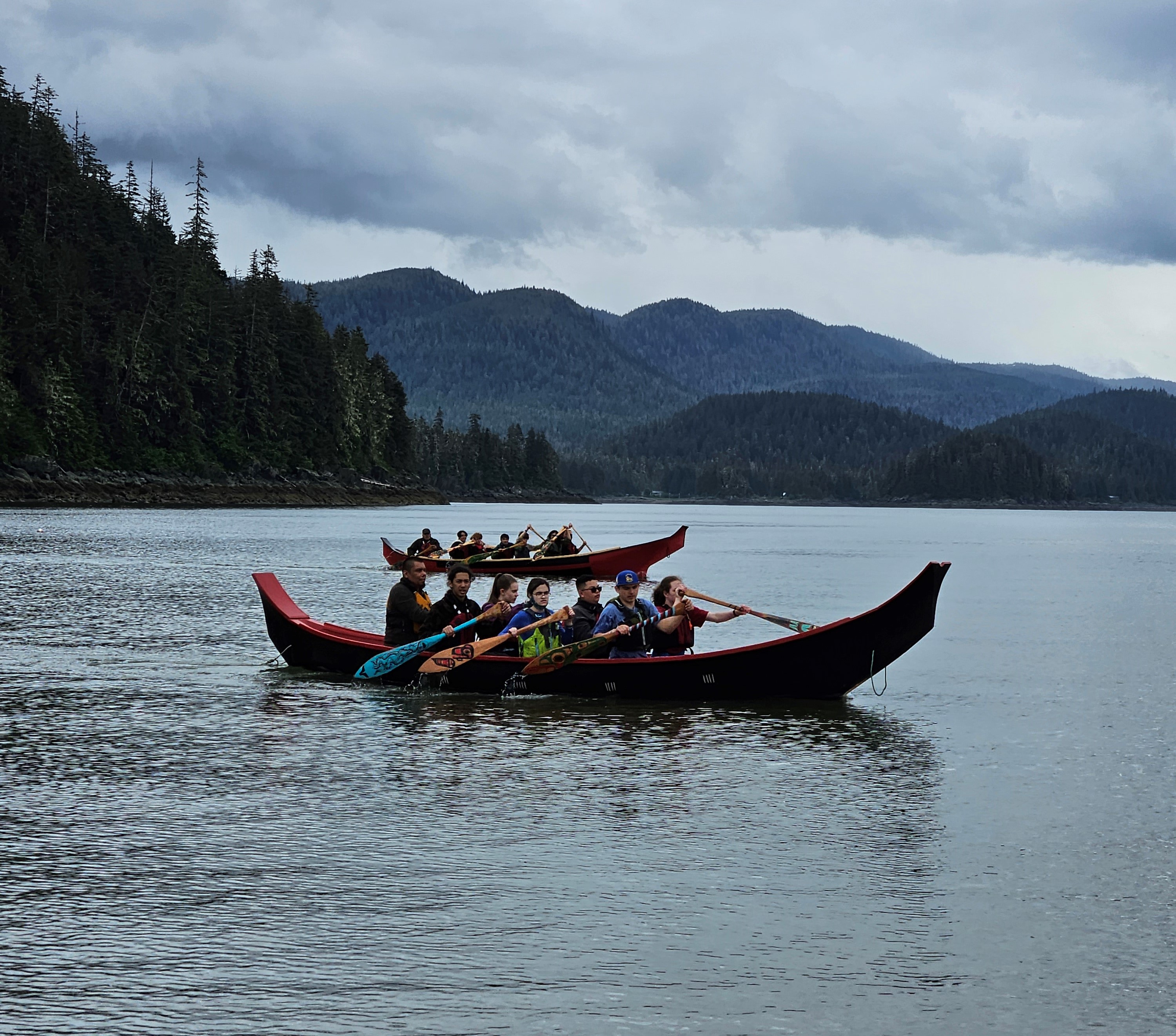 A group of people canoeing in two separate long canoes, one in the foreground and one in the background, on a lake surrounded by mountains.