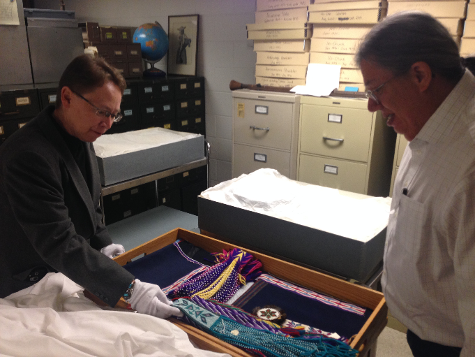 Two men in an office space wear gloves to handle a small box containing several bright colored artifacts.