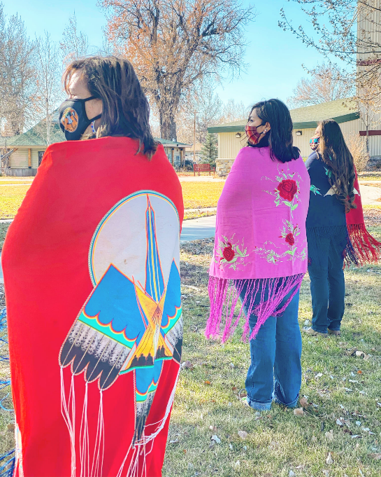 Three women wearing face masks stand outside in a line to show the back of the bright colored red and pink shawls they are wearing.