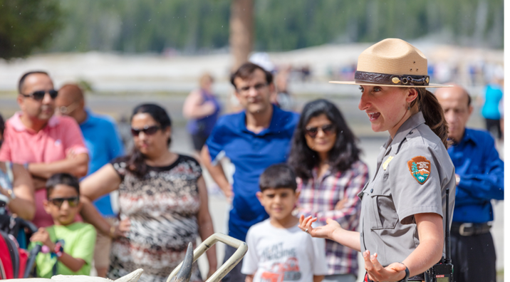 In the foreground to the right, a ranger, wearing a uniform and flat hat, speaks to members of the public in the background.