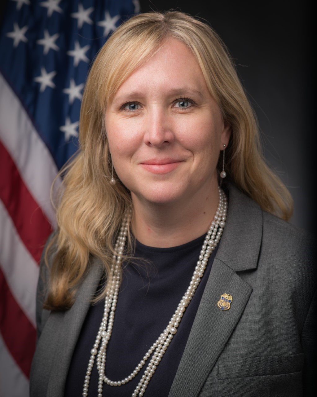 A middle-aged white woman with blonde hair in a grey suit accessorized with long pearl necklaces. Behind her is an American flag.