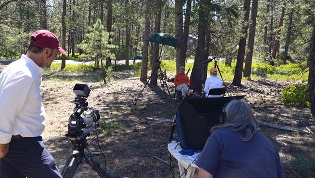 Two people film a tribal member being interviewed in the woods.